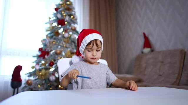 Beautiful baby boy in red cap sitting at desk. Toddler drawing with a blue pencil. Christmas tree at backdrop in blur.