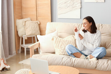 Young woman in warm sweater sitting on sofa at home - obrazy, fototapety, plakaty