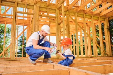 Father with toddler son building wooden frame house. Male builder teaching his son the technique of pounding nails with hammer on construction site, wearing helmets and blue overalls.