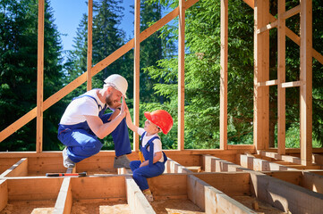 Father with toddler son constructing wooden frame house. Male builder giving high five to kid on...