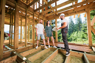 Man worker with detailed plan in hand showcases to young couple process of constructing wooden...