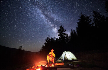 Night-time camping expedition in mountains, beneath stars. Male adventurer takes respite by bonfire...