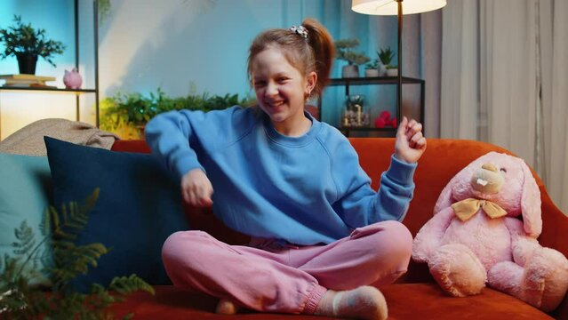 Trendy cheerful positive redhead girl having fun dancing, moving to rhythm, dabbing raising hands, making dub dance gesture. Teenager child kid at home in night evening play room sitting on couch