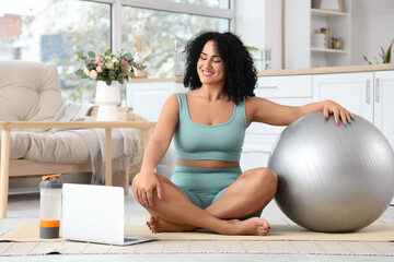Sporty adult woman with fitball and laptop sitting on yoga mat in kitchen