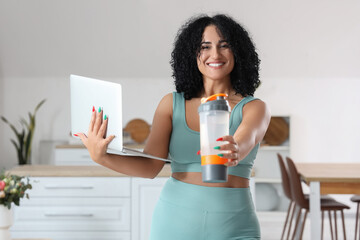 Sporty adult woman with modern laptop and bottle of water in kitchen