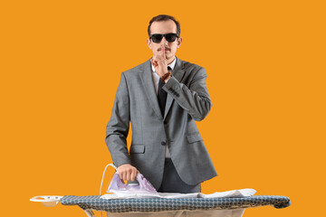 Young businessman ironing shirt on board and showing silence gesture against yellow background
