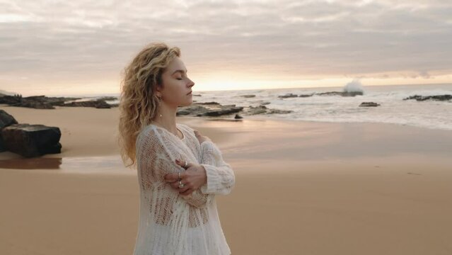 A young woman in white stands alone on a beach, hugging herself, and contemplating the ocean at sunset