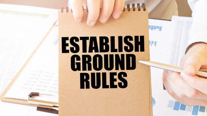 Text ESTABLISH GROUND RULES on brown paper notepad in businessman hands on the table with diagram....