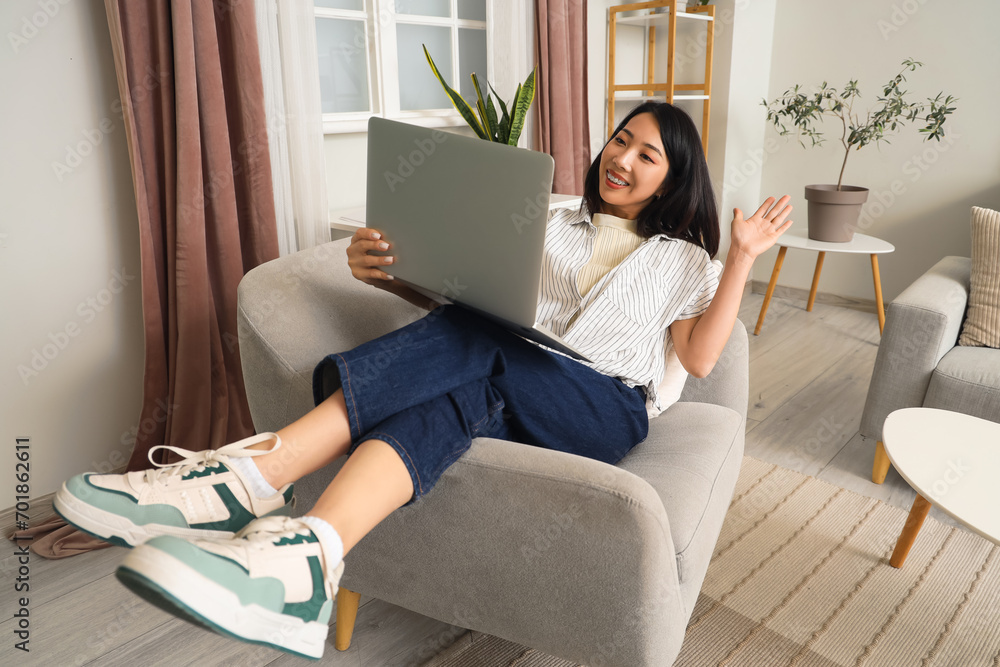 Poster Young Asian woman using laptop in armchair at home