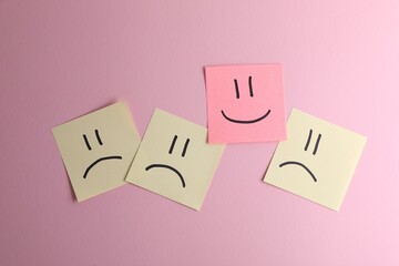 Choice concept. Sticky note with happy emoticon among beige papers with sad emojis on pink...