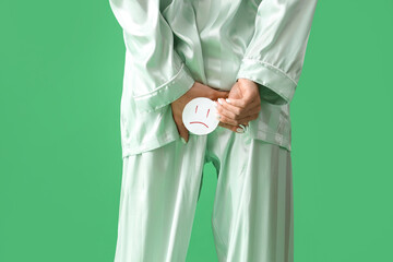Young woman in pajamas with hemorrhoids and sad smile on green background, back view
