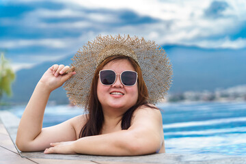Overweight young asian woman wearing orange swimsuit and straw beach sun hat relaxing in pool...
