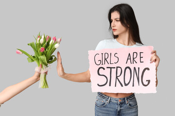 Young woman with GIRLS ARE STRONG sign rejecting bouquet of flowers on grey background. Feminism...