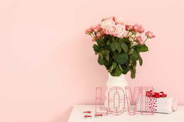 Vase with bouquet of roses and gift box on white cabinet near pink wall, closeup. Valentine's Day celebration