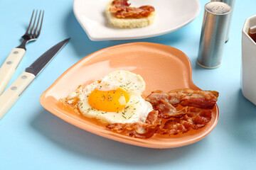 Plate with tasty bacon, heart made of fried egg and cutlery on blue background, closeup