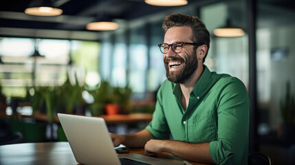 Cheerful man with a beard and glasses working on a laptop at a wooden desk - obrazy, fototapety, plakaty