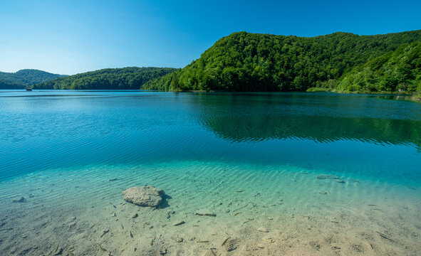 See im Nationalpark Plitvicer Seen, Kroatien. Flaches Ufer im Vordergrund, im Hintergrund H&uuml;gel mit B&auml;umen bei blauem Himmel im Sommer.