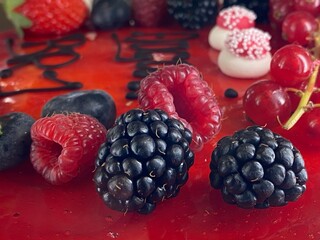 Cake decoration close up, blackberries in the forefront and other beautiful berries and sugar decoration on a vibrant red glaze. 