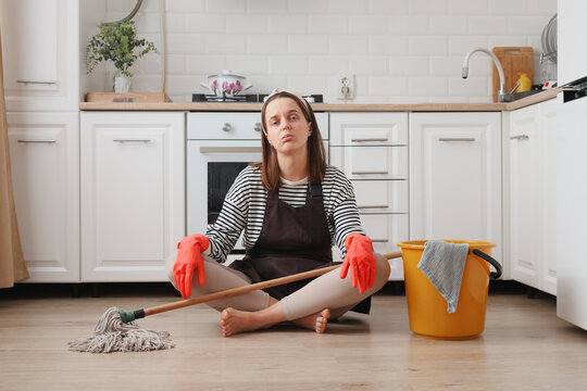 Upset woman wearing apron and rubber gloves housewife cleaning floor with steam mop in kitchen at home surrounded with cleaning stuff expressing negative emotions and fatigue