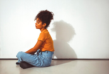 Calm pensive african american girl sitting in lotus pose on floor against white wall with shadow