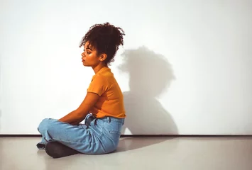 Poster Calm pensive african american girl sitting in lotus pose on floor against white wall with shadow © Dasha Petrenko