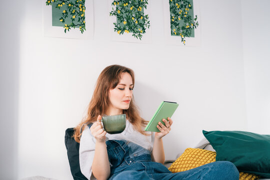 Young woman reading e-book on tablet reader in bed. Female relaxing with hot drink while reading an electronic book in cozy room. Mental health recovery, Personal growth. Hobby, Leisure time alone.