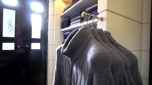 Panning over several men's gray wool sweaters on hangers in a men's clothing store with white walls and black doors. High quality and a large selection of warm clothes in a man's wardrobe