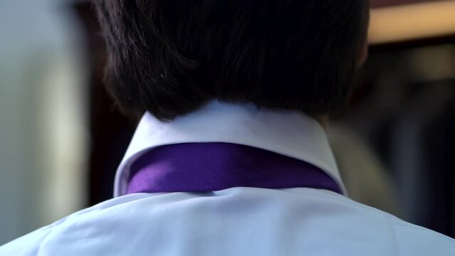 Close-up of the back of the head with thick black hair and the neck of an adult man wearing a white shirt and purple silk tie. Female staff serving a customer in a men's clothing store