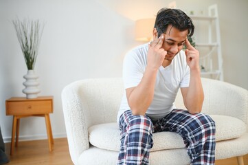 Portrait of young indian guy suffering from migraine at home, eastern man feeling unwell, touching his temples with closed eyes, suffering acute headache while sitting on couch