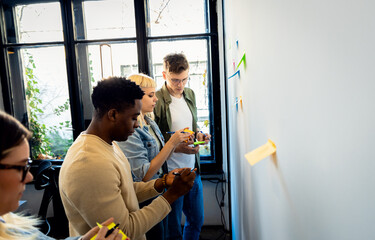 Diverse group of young business people working in office using colorful sticky notes on wall to...