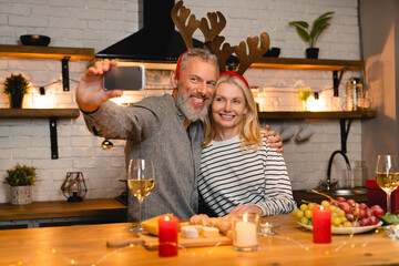Smiling happy mature couple taking selfie during Christmas dinner in the kitchen wearing deer`s hat