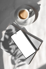 Minimal aesthetic business branding or social media template, mobile phone with empty white screen mockup, gray notebook, cup with milky coffee on marble table background with natural sunlight shadows