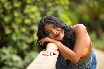 Naklejka premium Portrait of South American woman, young, beautiful, brunette, with colorful top and jeans leaning on a wooden railing with tender, pure, dreamy look. Concept of beauty, trend, ethnicity, diversity.