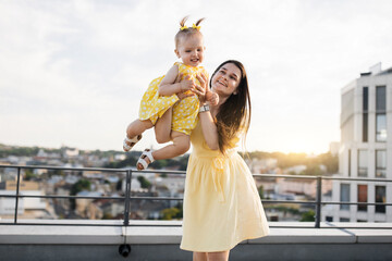 Happy young woman wearing yellow dress standing on roof of high-rise building and holding up little...