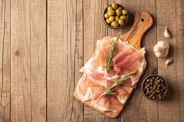 Thin slices of Italian prosciutto with green olives, spices and rosemary on wooden cutting broad....