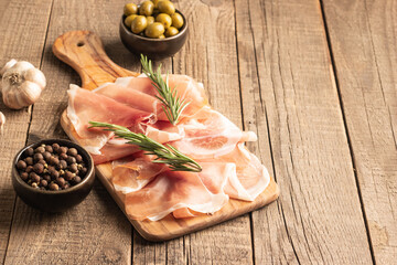 Thin slices of Italian prosciutto with green olives, spices and rosemary on wooden cutting broad. Raw dried meat.