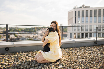 Fototapeta na wymiar Attractive lady with long dark hair sitting on ground made of small stones and holding black dachshund while hugging on roof of city building. Concept of best friends and love for pet.