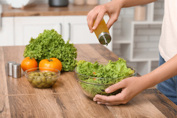 Woman adding olive oil into bowl with tasty salad at table in kitchen