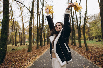 Attractive pretty woman walking outdoor, portrait of young lady in warm sunny autumn park season, fall, hold yellow orange red maple leaves, dressed leather wool fur jacket smiling having fun 

