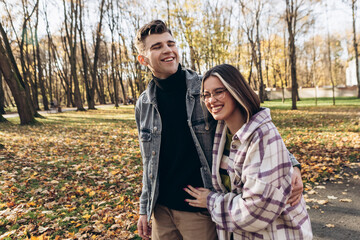 Heterosexual caucasian young loving couple walking outside in the city park in sunny weather, hugging smiling kissing laughing spending time together. Autumn, fall season, orange yellow red maple leav