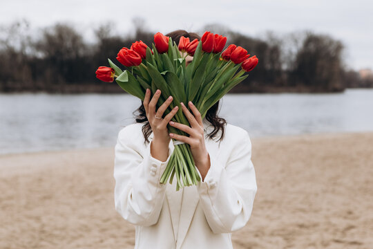 Attractive brunette woman walking on the beach shore in moody cloudy windy weather with bouquet of red tulips flowers, dressed in white suit jacket. International Women`s Day 8th March concept