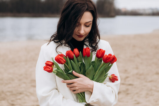 Attractive brunette woman walking on the beach shore in moody cloudy windy weather with bouquet of red tulips flowers, dressed in white suit jacket. International Women`s Day 8th March concept
