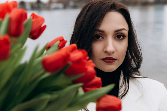 Attractive brunette woman walking on the beach shore in moody cloudy windy weather with bouquet of red tulips flowers, dressed in white suit jacket. International Women`s Day 8th March concept
