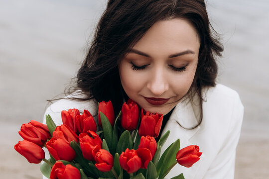 Attractive brunette woman walking on the beach shore in moody cloudy windy weather with bouquet of red tulips flowers, dressed in white suit jacket. International Women`s Day 8th March concept
