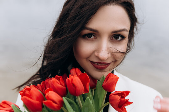 Attractive brunette woman walking on the beach shore in moody cloudy windy weather with bouquet of red tulips flowers, dressed in white suit jacket. International Women`s Day 8th March concept