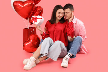 Young couple with gift and heart-shaped balloons sitting on pink background. Valentine's Day...