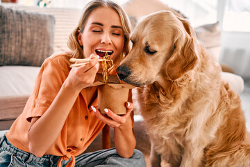 Togetherness with animal. Golden retriever eagerly licking paper box with chinese noodles being eating by young woman. Female pet owner sharing food with lovely furry friend at cozy apartment. - obrazy, fototapety, plakaty