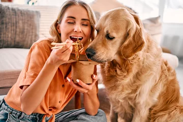 Fotobehang Togetherness with animal. Golden retriever eagerly licking paper box with chinese noodles being eating by young woman. Female pet owner sharing food with lovely furry friend at cozy apartment. © HBS