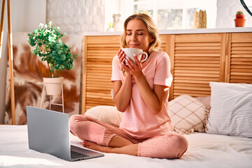 Relaxation at home. Carefree woman using laptop and drinking coffee while sitting on bed with...