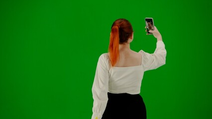 Portrait of attractive office girl on chroma key green screen. Woman in skirt walking and taking selfie on smartphone. Back view.